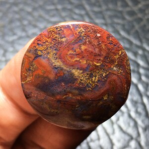 May include: A round, polished stone with a swirling pattern of red, orange, yellow, and blue hues. The stone's surface has a glossy finish, and the colors blend together in a natural, organic design. The stone is held in a hand.