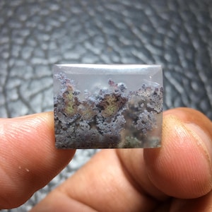 May include: A square, translucent gemstone with a landscape-like pattern. The design features a mix of gray, brown, and green formations, resembling a miniature natural scene. The stone is held between fingers.