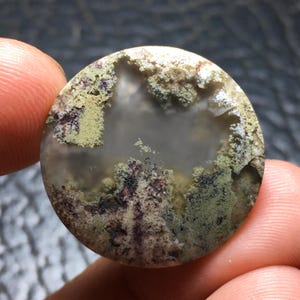 May include: A round, polished gemstone with a unique, natural pattern. The stone features a central, translucent area surrounded by moss-like formations in shades of green, brown, and purple. The overall effect is reminiscent of a miniature landscape.