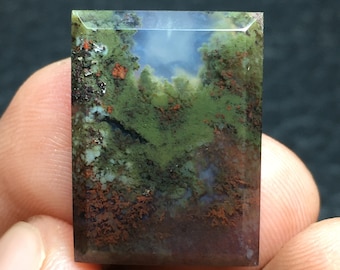 Può includere: Una pietra preziosa di agata muschiata rettangolare con un motivo a paesaggio. La pietra presenta inclusioni verdi, marroni e rosse, che ricordano una scena di foresta con un cielo blu. La superficie lucida esalta i colori e le trame naturali della pietra.