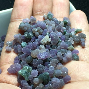 Puede incluir: Primer plano de un puñado de piedras de ágata de color púrpura, verde y gris. Las piedras son pequeñas y redondas, y tienen una superficie lisa y pulida.