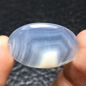 Puede incluir: Una piedra preciosa de ágata ovalada lisa, de color azul claro con bandas blancas y grises.