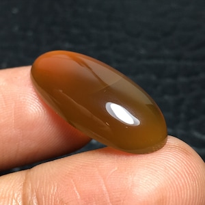 May include: A polished, oval-shaped gemstone with a gradient of colors, transitioning from a reddish-orange hue at one end to a deep brown at the other. The stone is held between two fingers, showcasing its smooth, reflective surface.