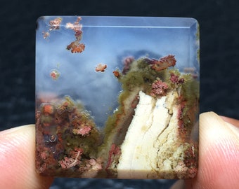 May include: A square, translucent agate cabochon with a white, layered, and textured pattern. The agate has a cloudy, blue-grey background with red and green inclusions.
