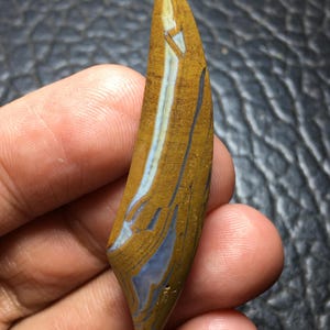 May include: A crescent-shaped polished gemstone cabochon featuring a smooth surface with distinct bands of mustard yellow, tan, and translucent light blue. The stone has a pointed top and bottom with a curved, elongated silhouette and natural patterns.