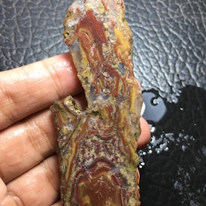 May include: A polished slab of agate stone featuring intricate, swirling patterns in shades of deep red, mustard yellow, and translucent white. The stone has an irregular, elongated shape with a rough, natural edge and a smooth, glossy surface.