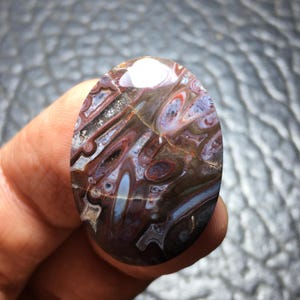 May include: An oval-shaped polished gemstone cabochon featuring a complex pattern of swirling brown, deep red, and translucent white bands. The stone displays a smooth, reflective surface with natural inclusions and distinct concentric ring formations.