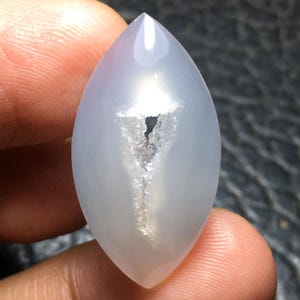May include: A marquise-shaped, translucent light blue chalcedony gemstone cabochon featuring a central, jagged, crystalline druzy inclusion that forms a distinct Y-shaped pattern. The stone has a smooth, polished surface with a soft luster.