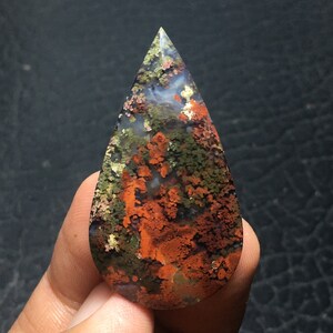 May include: A teardrop-shaped agate cabochon with a unique pattern of green, brown, and orange. The stone has a translucent quality and shows a variety of colors and patterns.