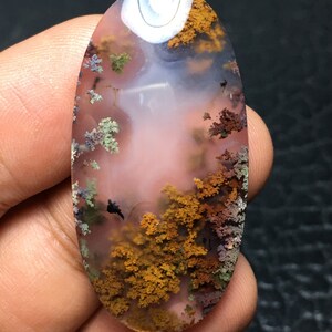 Puede incluir: Un cabujón de ágata pulido de forma ovalada con un patrón dendrítico. La ágata es translúcida con una base rosa y presenta dendritas marrones, verdes y negras.