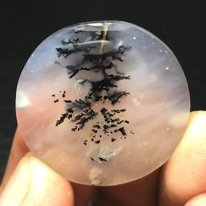 May include: A round, translucent, white agate cabochon with black dendritic inclusions. The agate has a cloudy, milky appearance with a hint of pink.