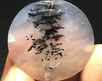 Könnte beinhalten: Ein runder, durchscheinender, weißer Achat-Cabochon mit schwarzen dendritischen Einschlüssen. Der Achat hat ein wolkiges, milchiges Aussehen mit einem Hauch von Rosa.