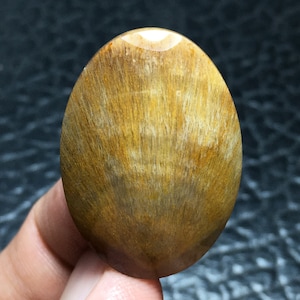 Könnte beinhalten: Oval geformter, polierter Edelstein mit goldbrauner Farbe und einem faserigen, strahlenförmigen Muster. Der Stein hat eine glatte, glänzende Oberfläche. Die natürliche Schönheit des Edelsteins wird durch das Licht hervorgehoben. Der Stein scheint in einer Hand gehalten zu werden.