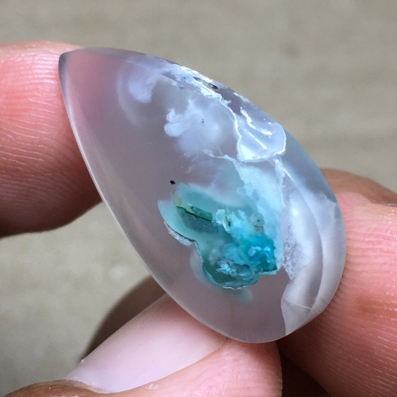 Peut inclure: Un cabochon en agate translucide en forme de larme avec des inclusions blanches et bleues. L'agate est de couleur gris clair avec des tourbillons blancs et un amas bleu-vert au centre.