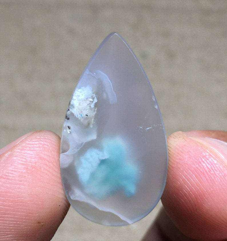 Peut inclure: Un cabochon d'agate en forme de larme avec un motif translucide blanc et bleu. L'agate a un aspect nuageux, laiteux avec un centre bleu-vert.
