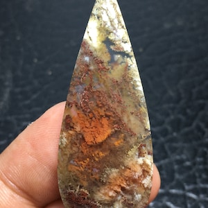 May include: A teardrop-shaped agate cabochon with a mix of white, brown, and orange colors. The stone has a unique pattern of dendrites and inclusions.