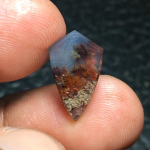 May include: A gemstone pendant with a unique landscape design. The top of the pendant is a light blue color, transitioning to a landscape of orange, brown, and green. The pendant is held between two fingers.