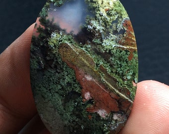 May include: Oval shaped agate cabochon with a green, brown, and white dendritic pattern. The agate has a translucent appearance with a cloudy white center.