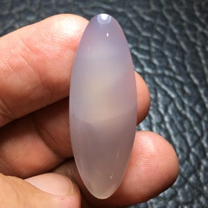 May include: A smooth, polished, marquise-shaped cabochon gemstone with a translucent, pale lavender-grey color. The stone features a soft, uniform surface with a subtle sheen and rounded edges, displaying a consistent light purple hue throughout.