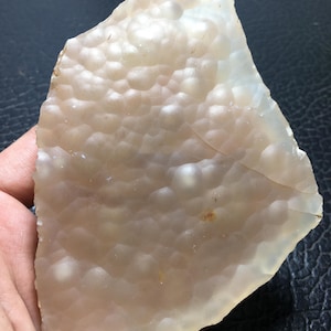 May include: A translucent, irregularly shaped stone with a pale peach hue. The surface has a textured, bubbly pattern, with areas of lighter and darker coloration. The stone is held in a hand, showcasing its natural form and unique characteristics.