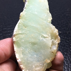 Può includere: Un esemplare di minerale grezzo verde chiaro, con una superficie ruvida e strutturata. La pietra ha una qualità traslucida con aree bianche e marroni. Il minerale è tenuto in una mano, mostrando la sua forma naturale e le variazioni di colore.