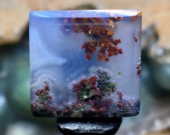 May include: A translucent, light blue agate cabochon with dendritic inclusions of brown and green. The agate has a cloudy, swirling pattern and is set on a black display stand.