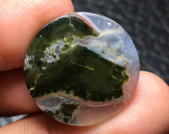 Puede incluir: Una gema redonda y pulida con un patrón único. La piedra presenta una mezcla de tonos verde oscuro, blanco y marrón, creando una apariencia natural y orgánica. La gema se sostiene entre los dedos, mostrando su tamaño y detalle.