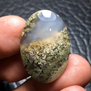 Può includere: Una gemma di agata muschiata di forma ovale caratterizzata da una sezione superiore traslucida bianco latte e una sezione inferiore densa e strutturata, ricca di complesse inclusioni simili a muschio nei toni del verde bosco e marrone.