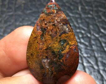 May include: A teardrop-shaped gemstone with a polished surface. The stone features a blend of earthy colors, including shades of brown, orange, yellow, and green. The gemstone's unique pattern and texture create a natural, organic appearance. The stone is held in a hand.