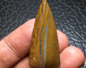 May include: A polished, triangular gemstone with a pointed tip. The stone features a blend of golden-brown and tan hues, with streaks of blue and gray running through it. The gemstone is held in a hand.