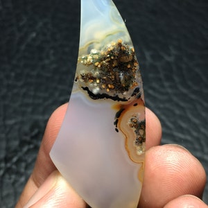 May include: A white agate cabochon with brown, black, and green inclusions. The agate is shaped like a teardrop and has a smooth, polished surface.