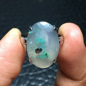 Pode incluir: Um anel de prata com uma gema oval branca com inclusões verdes e pretas. O anel é engastado com garras.