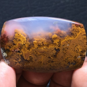 May include: A polished agate cabochon with a cloudy white center and a brown, yellow, and red landscape pattern.