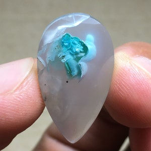 Peut inclure: Un cabochon d'agate en forme de larme avec un fond gris clair et une inclusion bleu-vert. L'inclusion est un amas de cristaux de forme irr&eacute;guli&egrave;re et rugueuse.