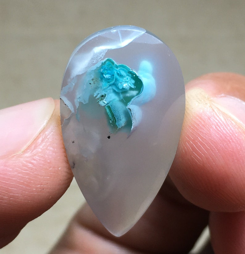 Peut inclure: Un cabochon d'agate en forme de larme avec un fond gris clair et une inclusion bleu-vert. L'inclusion est un amas de cristaux de forme irr&eacute;guli&egrave;re et rugueuse.