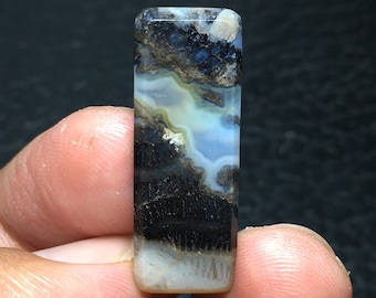 May include: A rectangular, polished agate cabochon with a white, blue, and black pattern. The agate has a layered, banded appearance with a white centre and black dendritic inclusions.