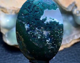 May include: A green moss agate cabochon with white and grey inclusions. The stone is oval shaped and has a polished surface.