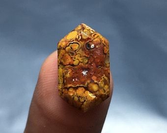Könnte beinhalten: Ein gelber, orangefarbener und brauner Achat-Cabochon mit einem einzigartigen, wirbelnden Muster. Der Edelstein ist in eine sechseckige Form geschnitten und hat eine glatte, polierte Oberfläche.