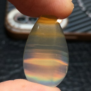 Puede incluir: Una piedra preciosa en forma de lágrima, iridiscente con un brillo arcoíris y una apariencia translúcida y en capas.