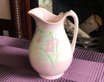 May include: A pale pink ceramic jug with a floral pattern of pink and green flowers. The jug has a curved handle and a spout. It rests on a purple striped tablecloth, with a fireplace visible in the background.