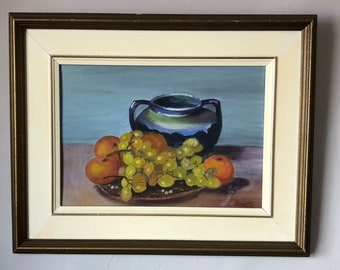 Könnte beinhalten: Ölgemälde in einem goldenen Rahmen mit einem Stillleben mit Obst und einer Vase. Das Gemälde zeigt eine dunkelblaue Vase mit grünem Rand, Orangen und grüne Trauben auf einem Teller. Der Hintergrund ist hellblau.