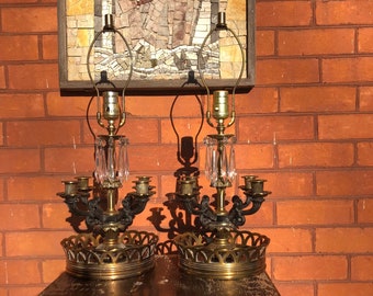 May include: Two antique brass lamps with crystal accents and ornate candle holders. The lamps sit on a wooden table in front of a brick wall and a mosaic artwork. The lamps have a vintage aesthetic.