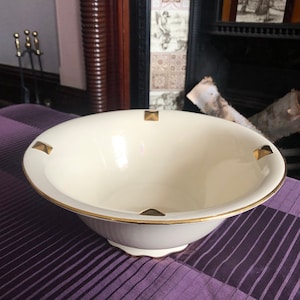 May include: Cream-coloured ceramic bowl with a gold rim and gold square accents. The bowl has a fluted exterior and sits on a decorative base. The bowl is on a purple tablecloth.
