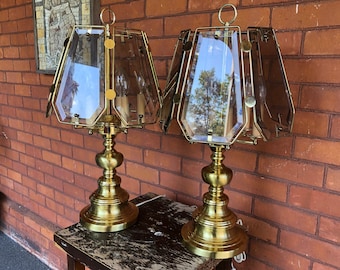 May include: Two vintage brass table lamps with geometric shades. The lamps have a classic design with a brass base and a clear, faceted shade. The shades are made of clear glass and are held together by brass frames.