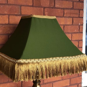 May include: A green lampshade with gold trim and fringe. The lampshade is square and has a gold trim around the top and bottom edges. The fringe is a light gold color and hangs down from the bottom edge.