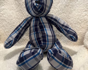 Pode incluir: Um urso de pelúcia feito à mão com tecido xadrez azul, branco e azul marinho. O urso tem um nariz preto e olhos pretos pequenos. O urso está sentado em uma superfície branca e fofa.