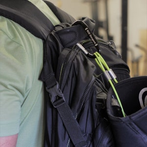 May include: A black backpack with a green strap and a carabiner clip attached to the strap. The carabiner has the text "WOD Hangers" printed on it.