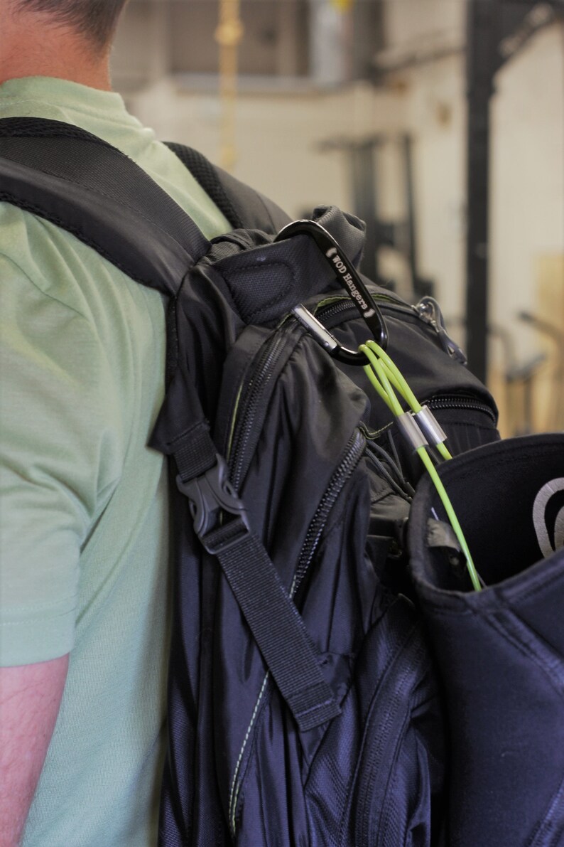 May include: A black backpack with a green strap and a carabiner clip attached to the strap. The carabiner has the text "WOD Hangers" printed on it.