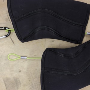 May include: Two black knee pads with green straps and silver clips. The straps are attached to the knee pads with a carabiner clip. The knee pads are made of a soft, flexible material.