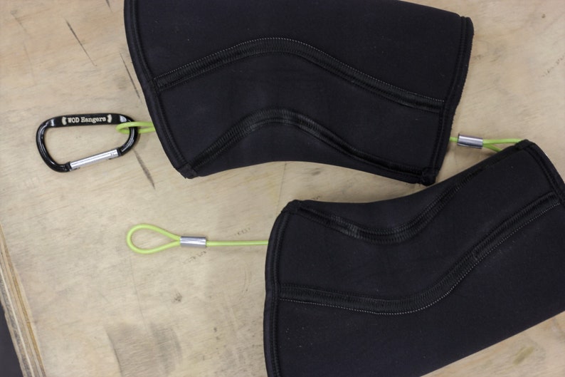 May include: Two black knee pads with green straps and silver clips. The straps are attached to the knee pads with a carabiner clip. The knee pads are made of a soft, flexible material.